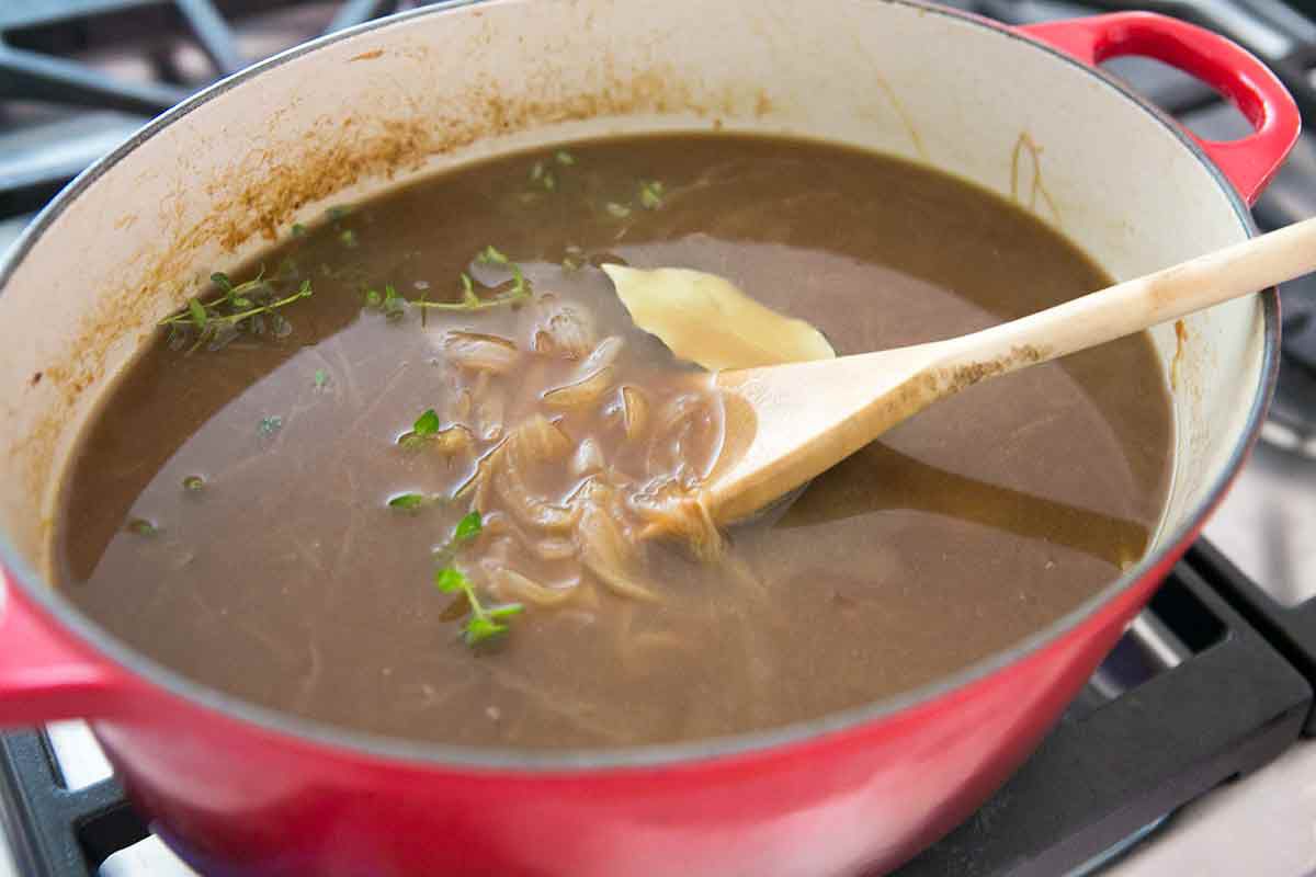Stock and herbs added to cooking onions in a pot