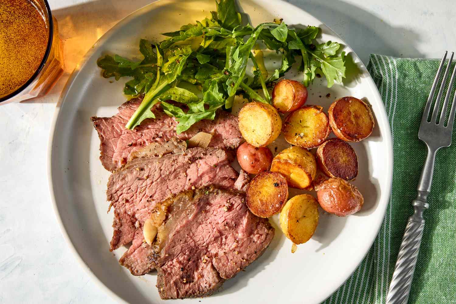 Roast beef slices, small roasted potatoes, and leafy greens served on a plate with a fork and napkin nearby