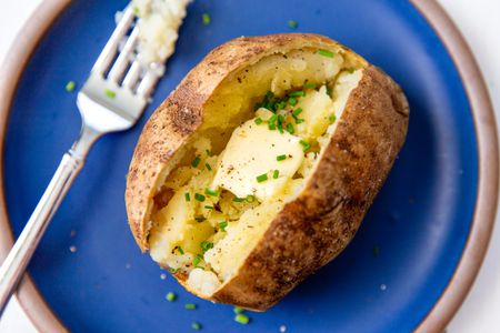 baked jacket potato cut open length wise and filled with butter and chives, on a blue pate with a fork on the left side