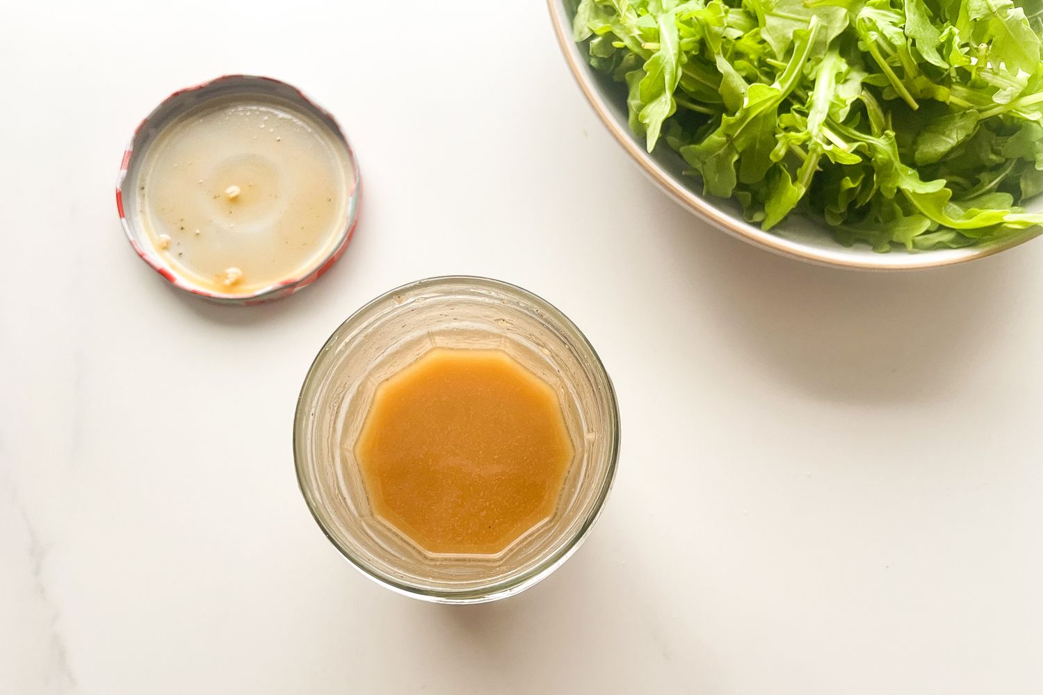 A jar of salad dressing next to a bowl of greens on a light surface