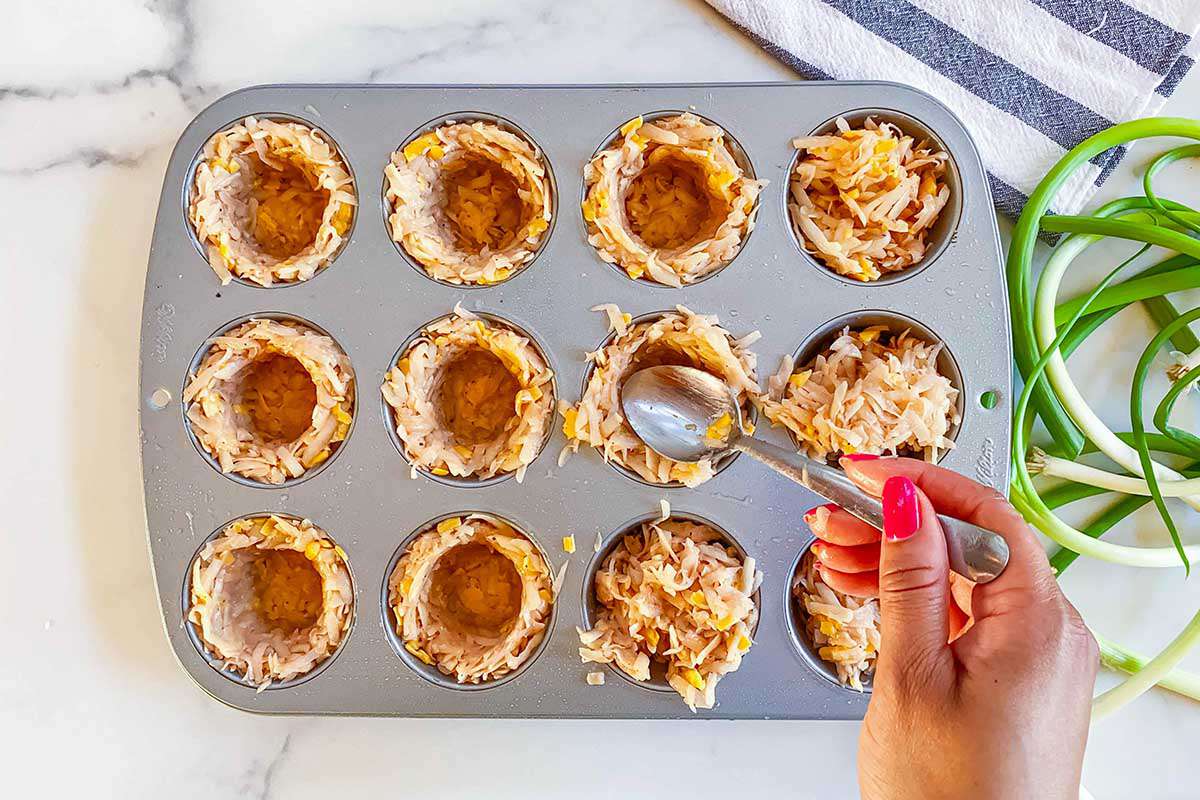 Hash brown egg bite crusts are being made by spooning shredded hashbrowns into the muffin tin cavity. Scallions and a striped linen are visible to the right.