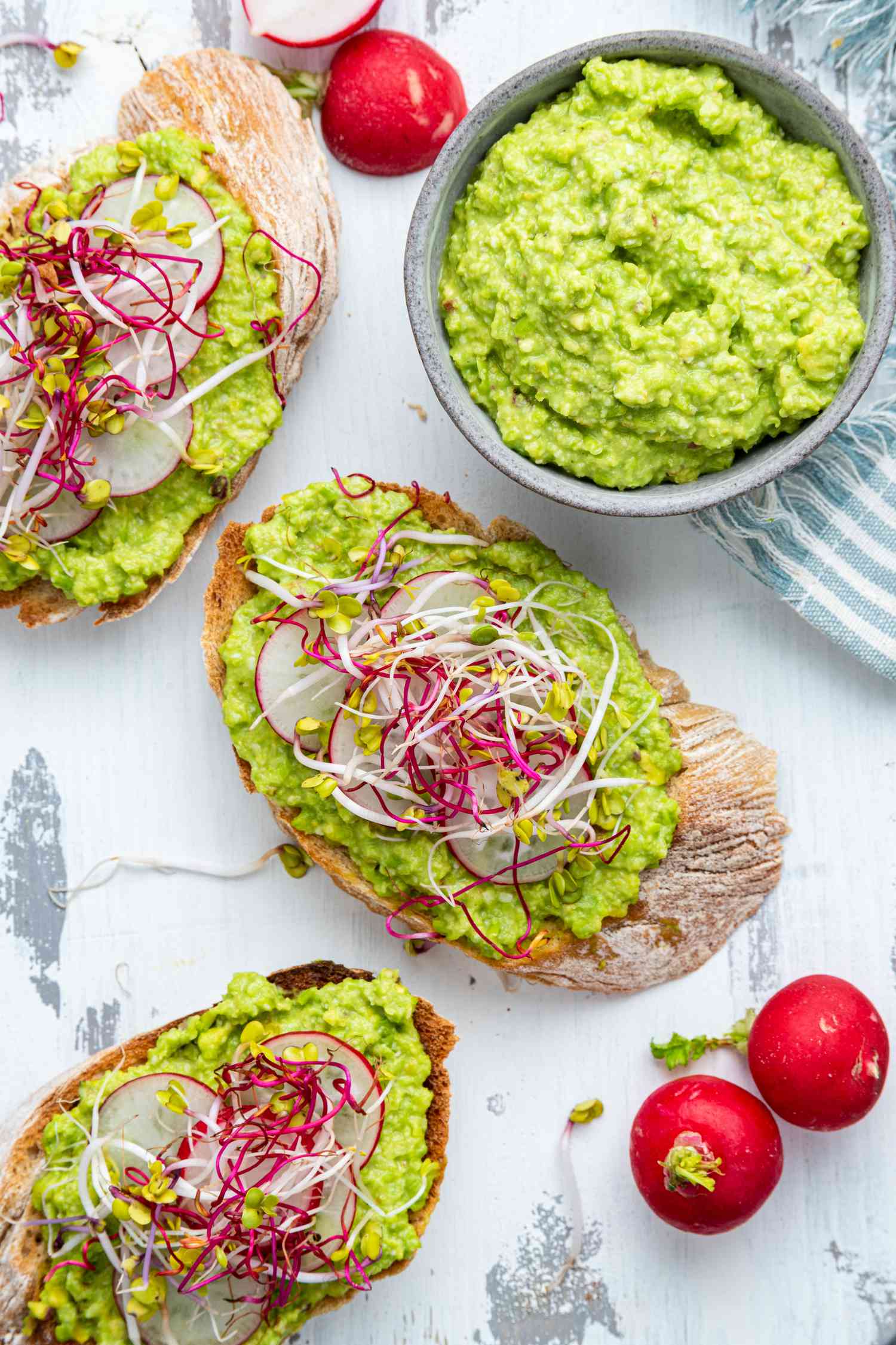 Slices of Toasted Smeared With Pea Pesto and Topped With Sliced Radishes and Microgreens, and in the Surroundings, Whole Red Radishes, a Bowl of Pea Pesto, and a Blue and White Stripped Kitchen Towel 