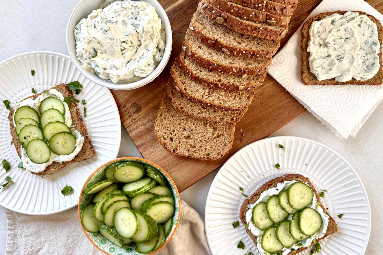 An assortment of sandwiches with cucumber slices on rye bread accompanied by a bowl of cucumber slices and spreadable topping on plates