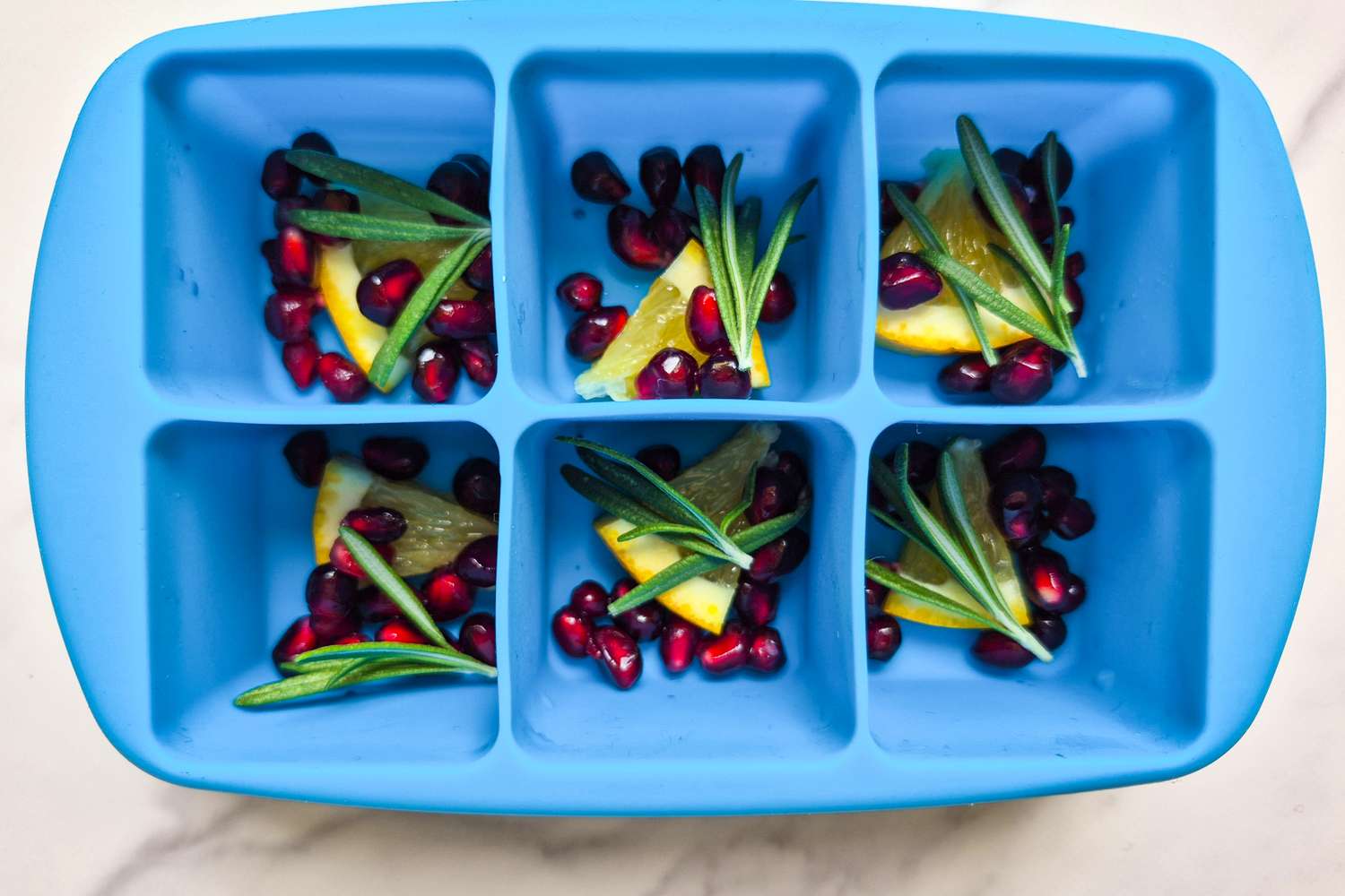 Blue silicone ice cube tray with compartments containing pomegranate seeds citrus slices and rosemary sprigs