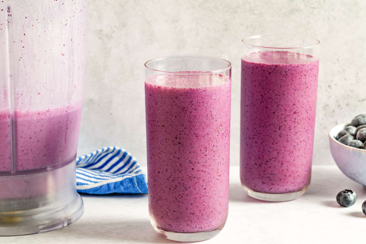 Two glasses of a blueberry smoothie next to a blender and bowl of blueberries