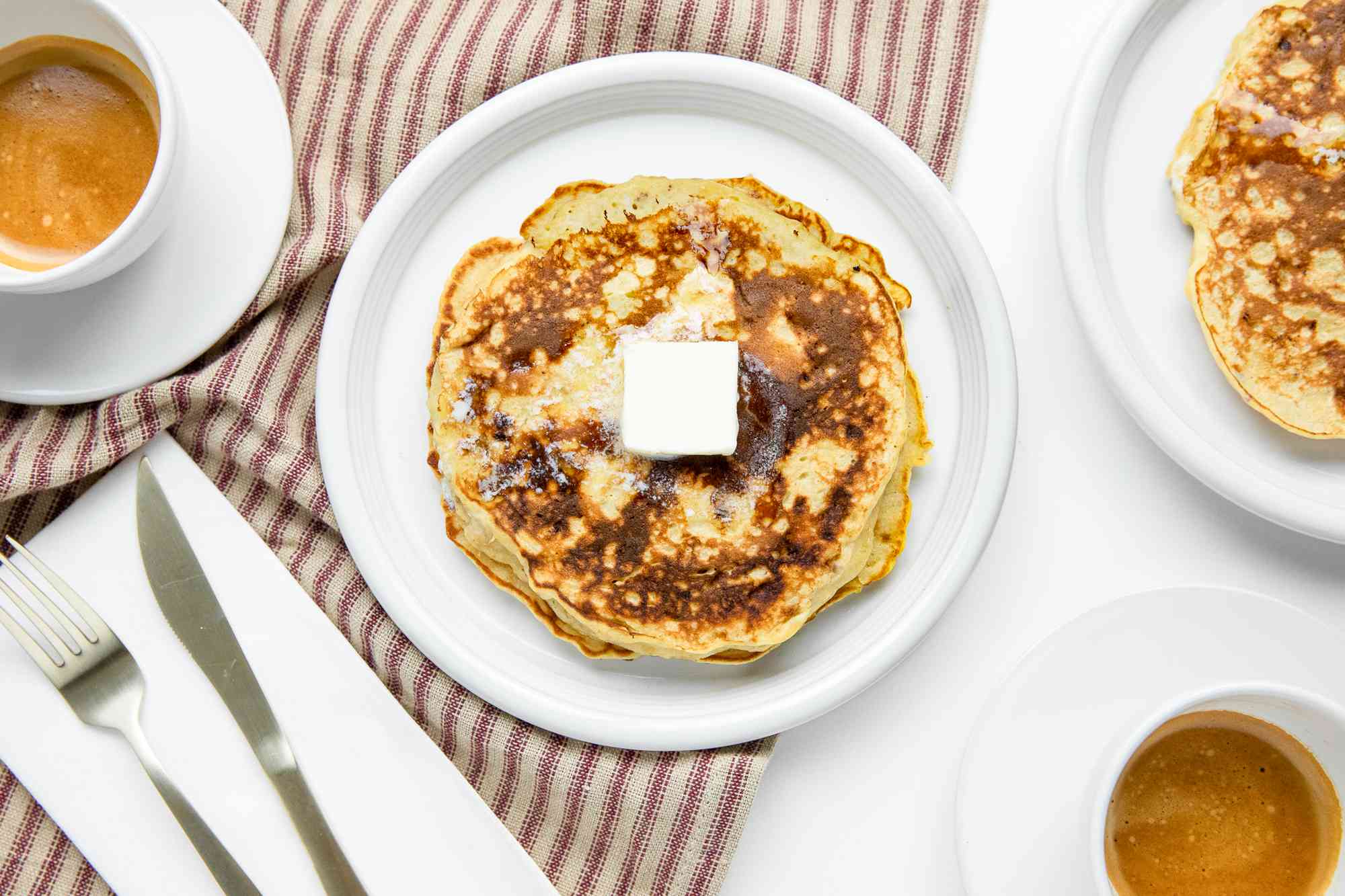Plate with a Stack of 3-Ingredient Banana Pancakes with Butter, Surrounded by a Plate with More Pancakes, Cups of of Coffee, and Utensils on a Table Napkin