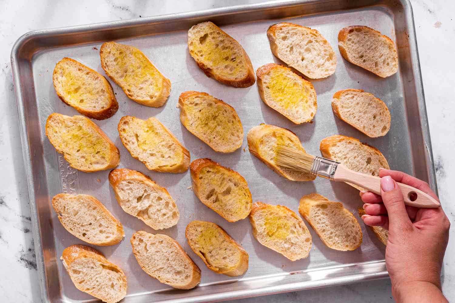 Olive oil brushed onto baguette slices on a baking sheet for bruschetta with tomato and basil recipe