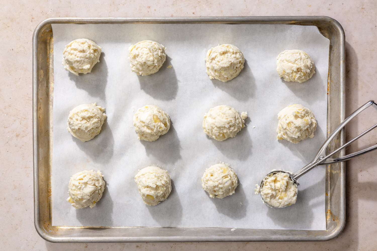 Overhead view of a using a cookie scoop to form the biscuits and add to parchment paper on a metal baking sheet for Mashed Potato Biscuits recipe