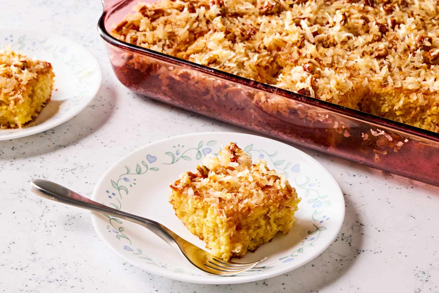 Angled view of a glass baking dish of pineapple coconut cake next to two small plates with slices