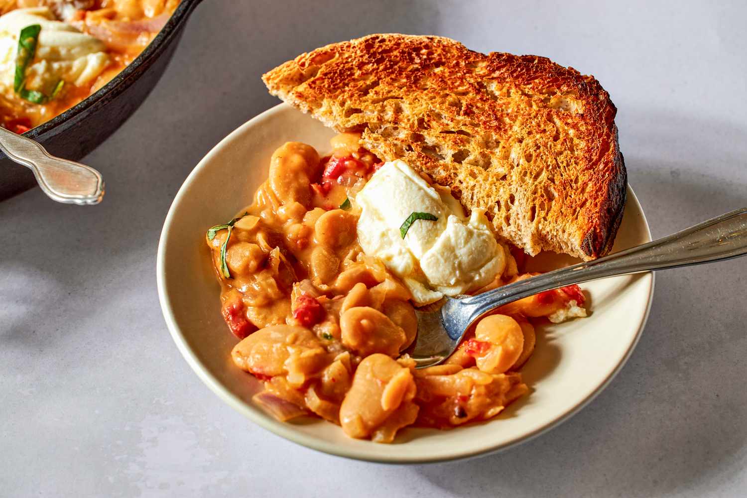 Side view closeup of a serving of Tuscan butter beans on a plate, with a dollop of ricotta on top and a slice of toasted bread on the side
