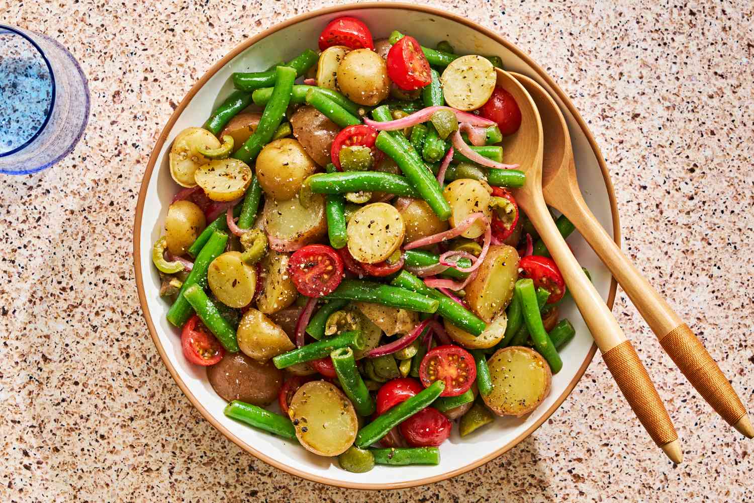 Sicilian potato salad in a bowl with serving utensils and next to a glass of water