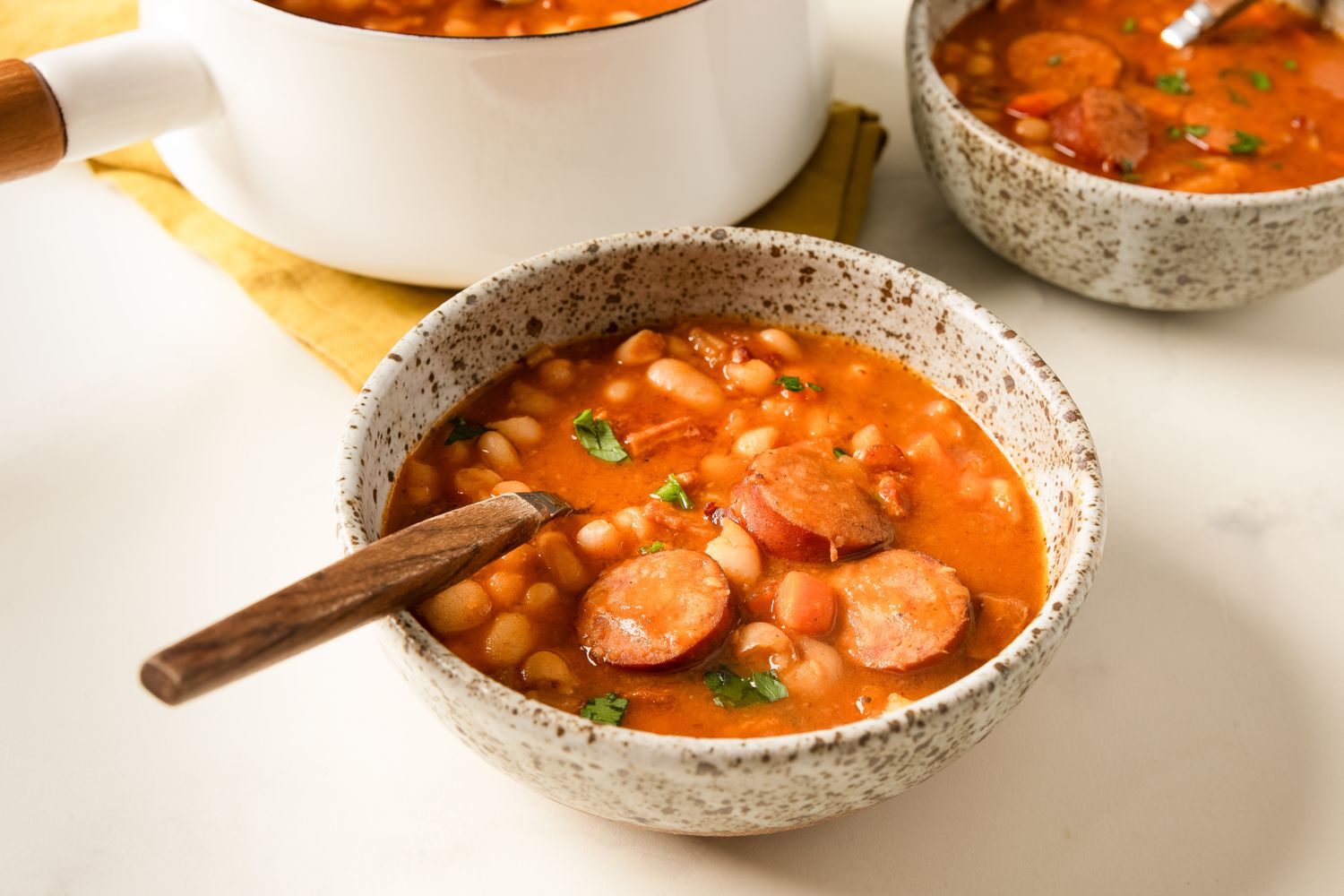 Bowl of bean and sausage stew with a spoon bread and other containers with the same dish in the background