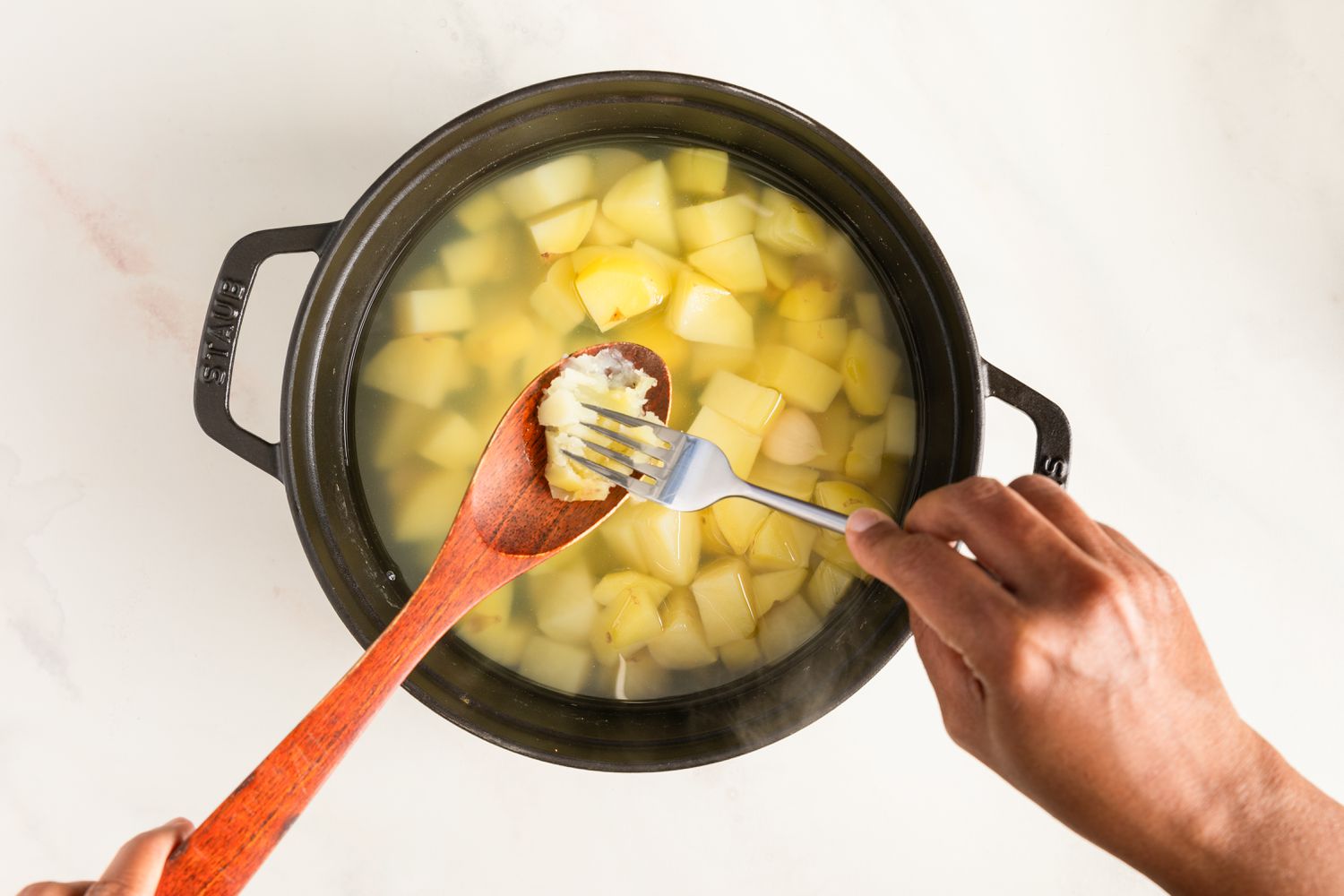 Boiled potatoes in a pot with one on a wooden spoon being mashed with a fork to test doneness
