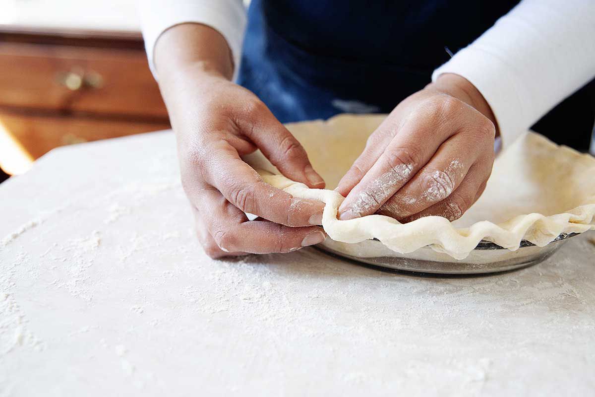 Woman in blue apron crimping pie dough.