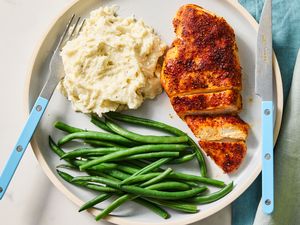 A plate with seasoned cooked chicken breast mashed potatoes and green beans accompanied by a fork and knife