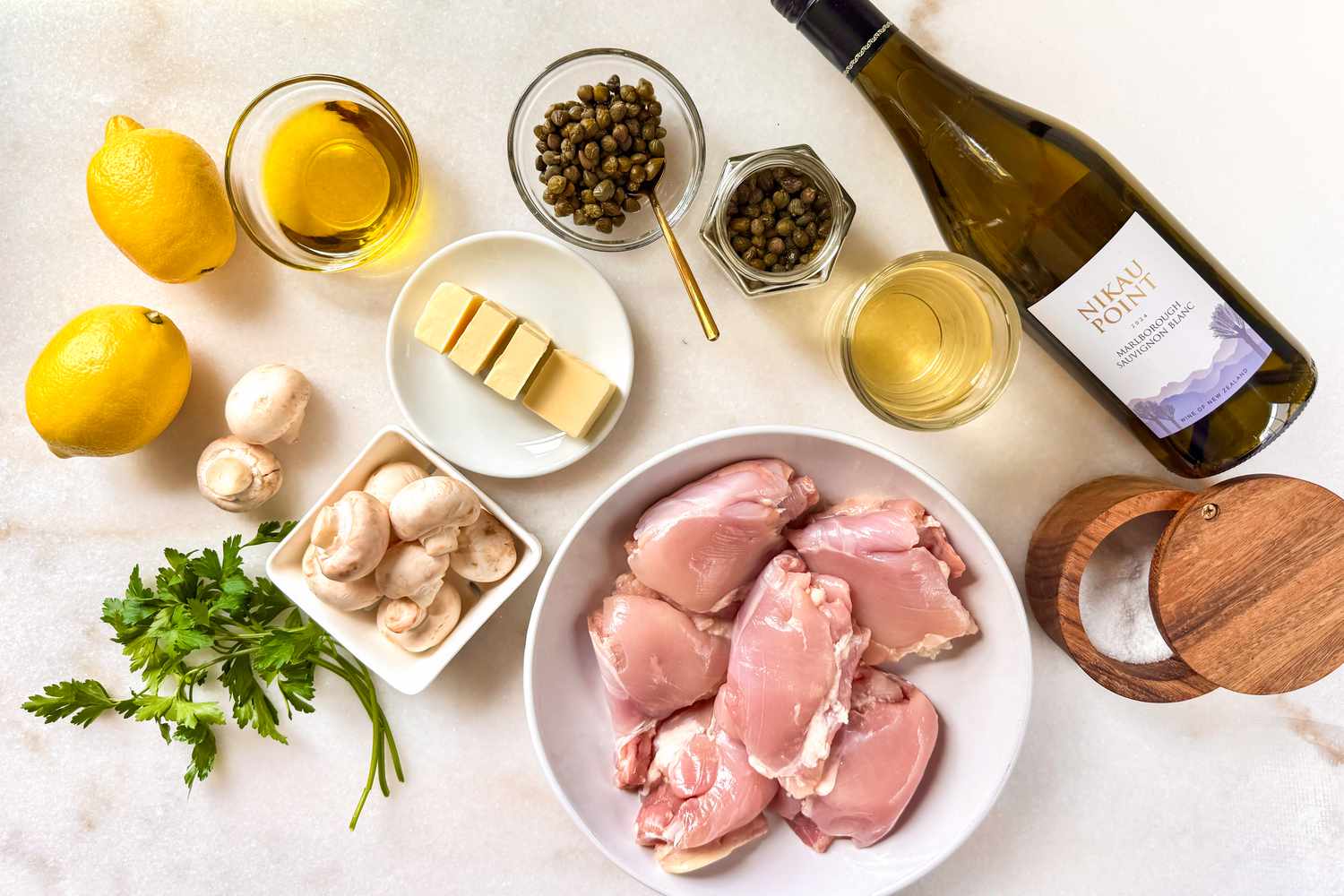 Ingredients for Alton Brown's chicken piccata recipe, including raw chicken, lemons, butter, mushrooms, capers, parsley, oil, wine, and seasonings