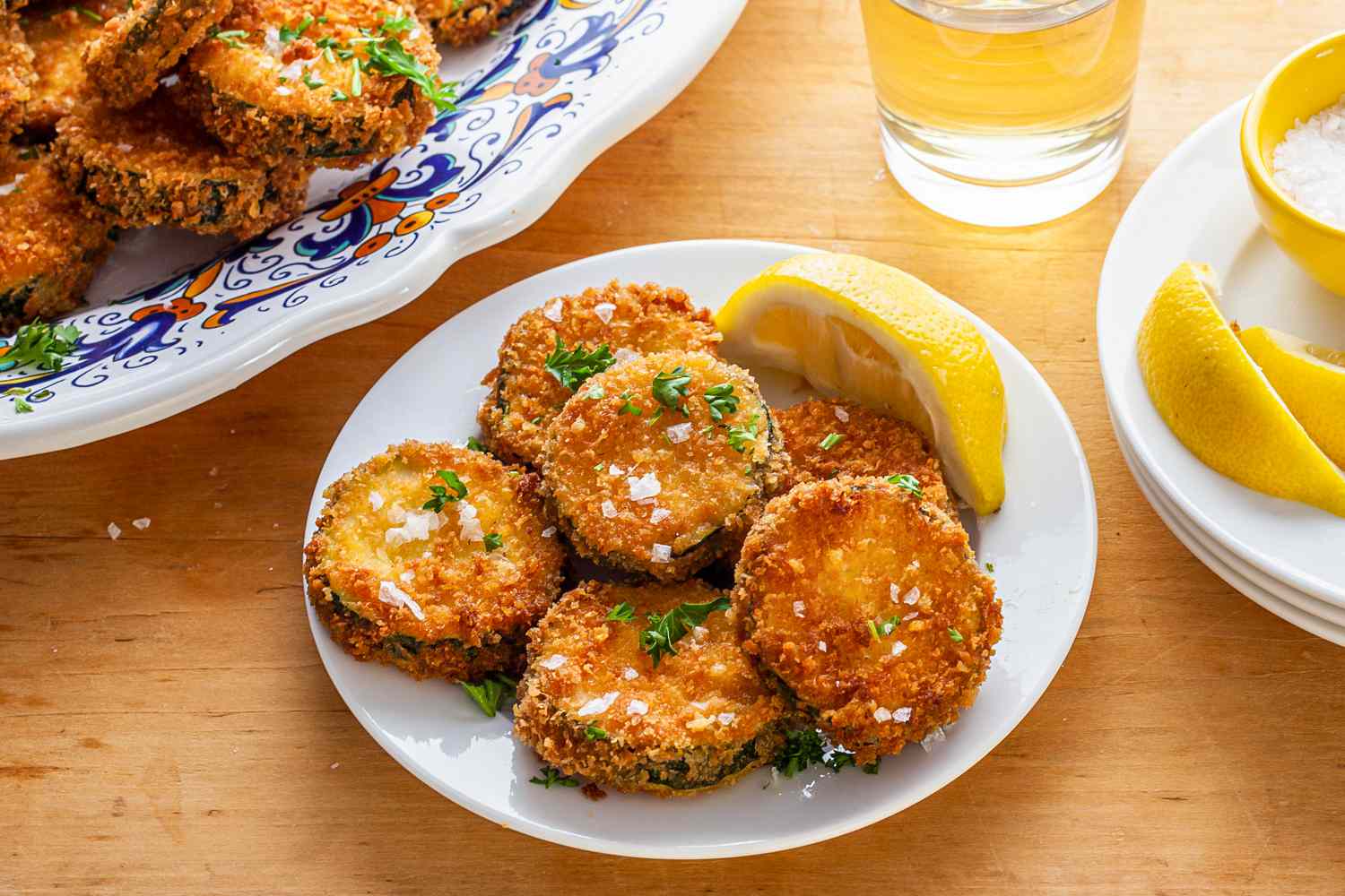 Plate of Fried Zucchini with a Lemon Wedge Next to Small Bowl of Salt, a Platter with More Fried Zucchini, and a Glass