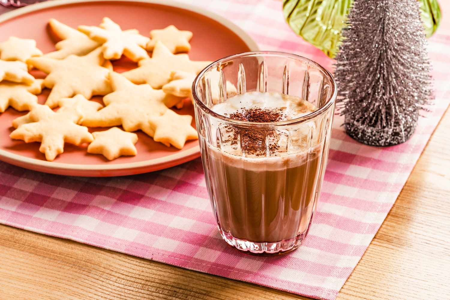 A glass of eggnog topped with grated chocolate beside a plate of starshaped cookies on a checkered tablecloth