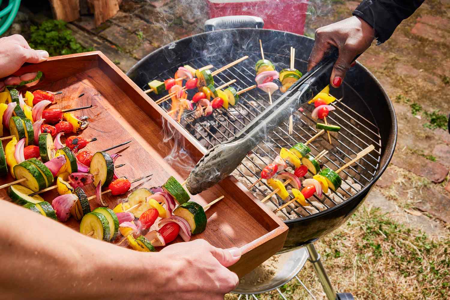 Vegetable skewers being placed on a barbecue grill with tongs by a pair of hands