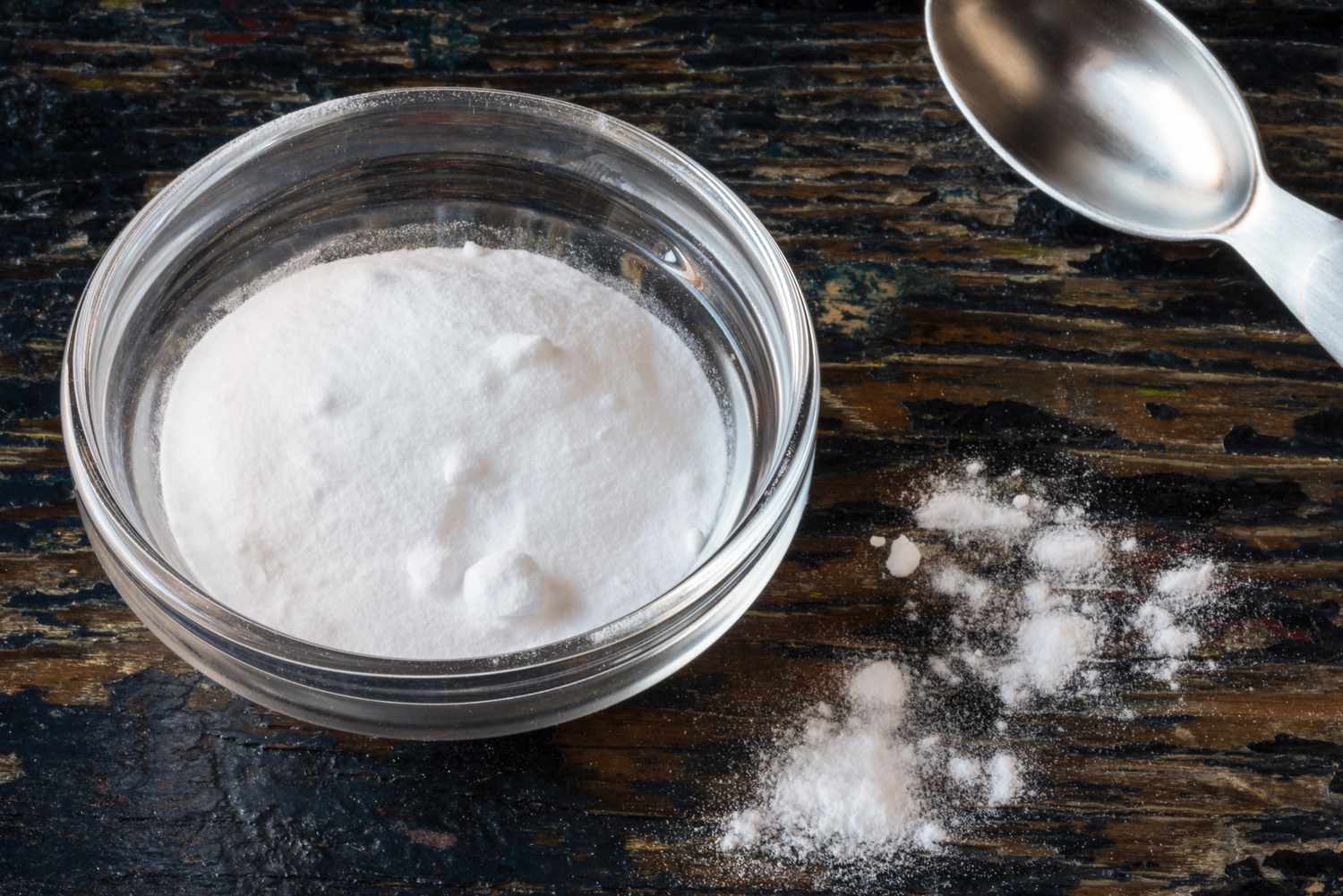 A small glass bowl of baking powder with a measuring spoon on a wood surface
