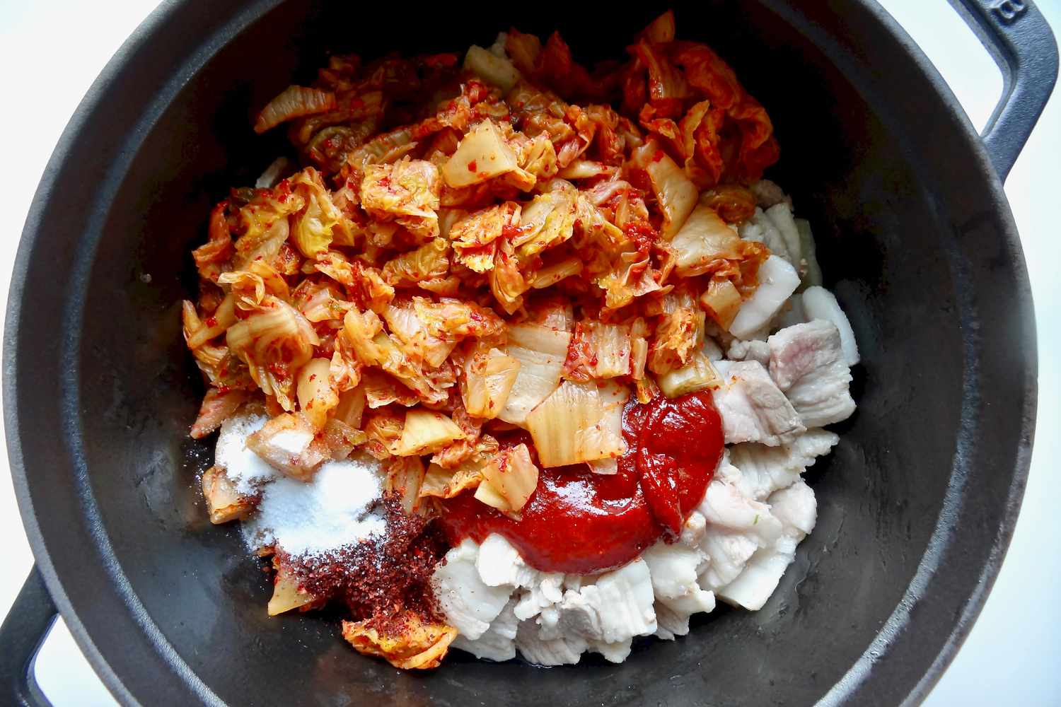 Adding ingredients to a dutch oven to make spicy kimchi and pork belly stew