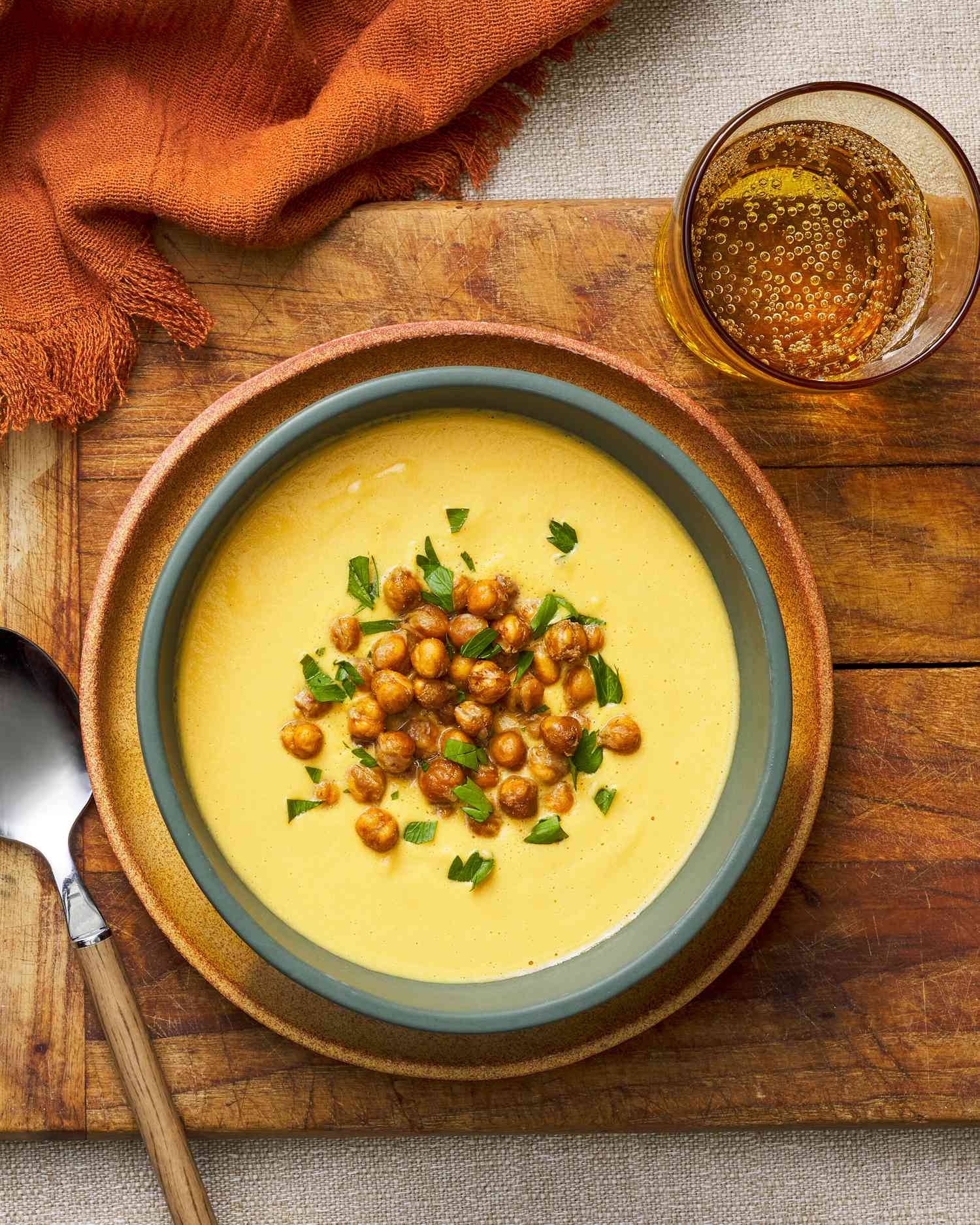 Bowl of hummus soup at a rustic table setting (bowl sitting on a wooden tray next to a glass of sparkling water, a spoon, and a burnt orange kitchen towel)
