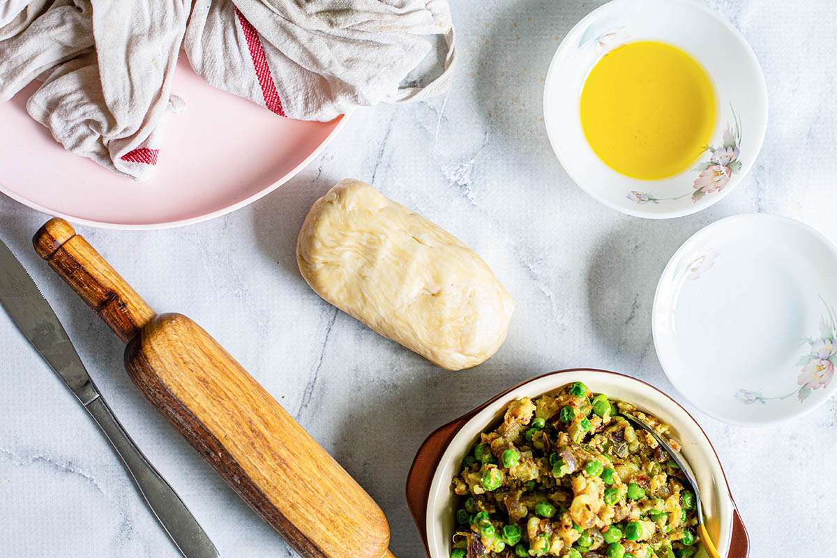 Bowl with vegetable samosa filling, a white bowl with ghee, a rolling pin, and a pink plate with a towel.