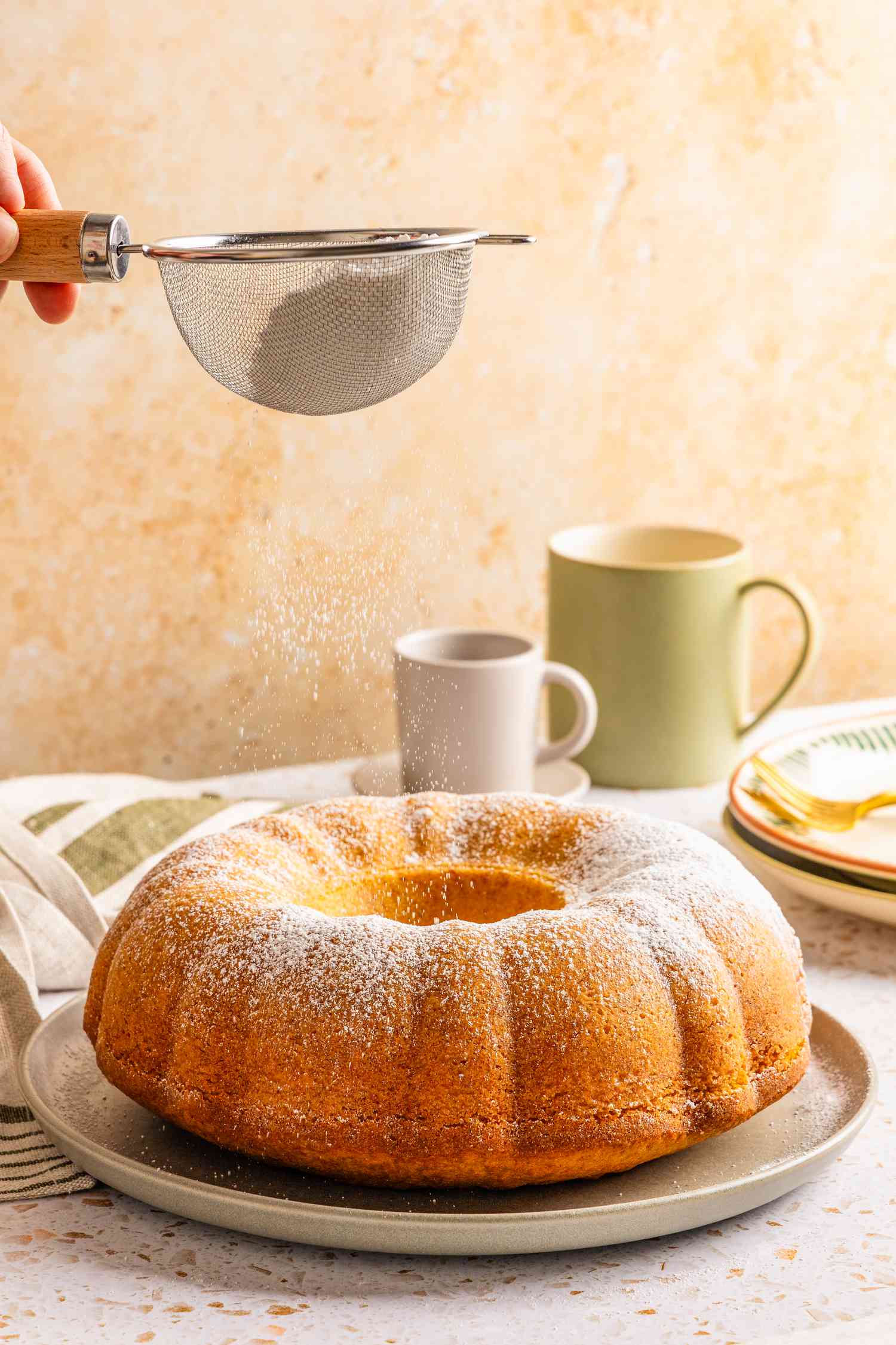 powdered sugar dusted onto ciambellone on a platter at a table setting with mugs, stack of plates, and table napkins