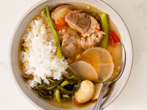 A bowl of pork sinigang served with rice and vegetables including radish green beans and eggplant with a spoon on the side