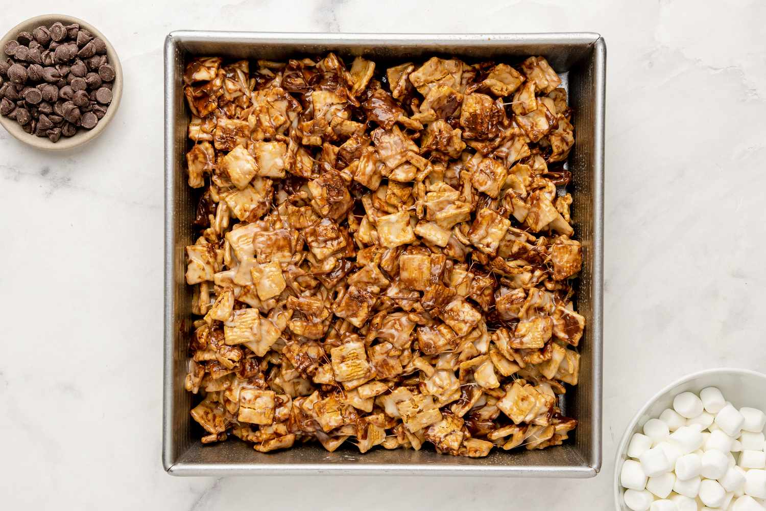 S’mores Cereal Mix in Baking Pan Surrounded by Bowls of Chocolate Chips and Marshmallows