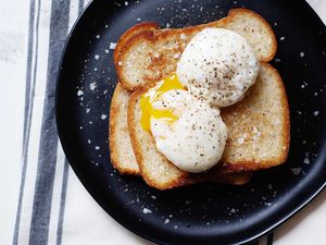 Two poached eggs on top of two pieces of stacked toast. One egg has a slightly runny yolk. Salt and pepper are sprinkled on the eggs, toast and black plate.