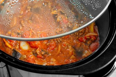 A slow cooker with a tomatobased stew visible through a glass lid containing meat onions and other vegetables