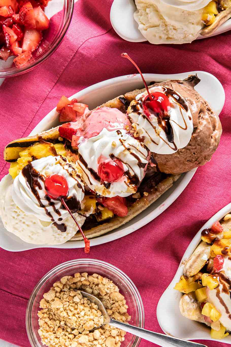 Grilled Banana Split Topped With Chocolate Sauce, Peanuts, Whipped Cream, and Three Maraschino Cherries in a Banana Split Dish, and in the Surroundings, Two More Banana Splits in Banana Split Dishes, a Bowl With Diced Strawberries, and a Bowl of Chopped Peanuts With a Spoon