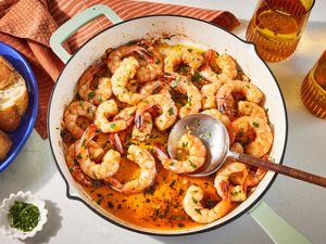 spanish garlic shrimp in a dutch oven at a table setting with forks on a table napkin, glasses of beer, and a serving of spanish garlic shrimp with baguette slices in a bowl