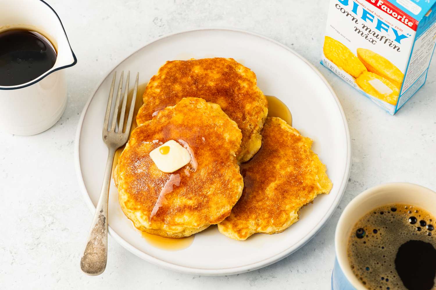 Jiffy Pancakes topped with syrup and a slab of butter on a plate at a table setting wtih a container of maple syrup, a box of jiffy muffin mix, and a mug of coffee