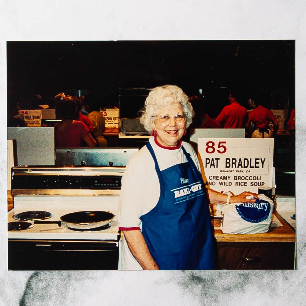 A person in an apron next to a display for creamy broccoli and wild rice soup at a cooking event