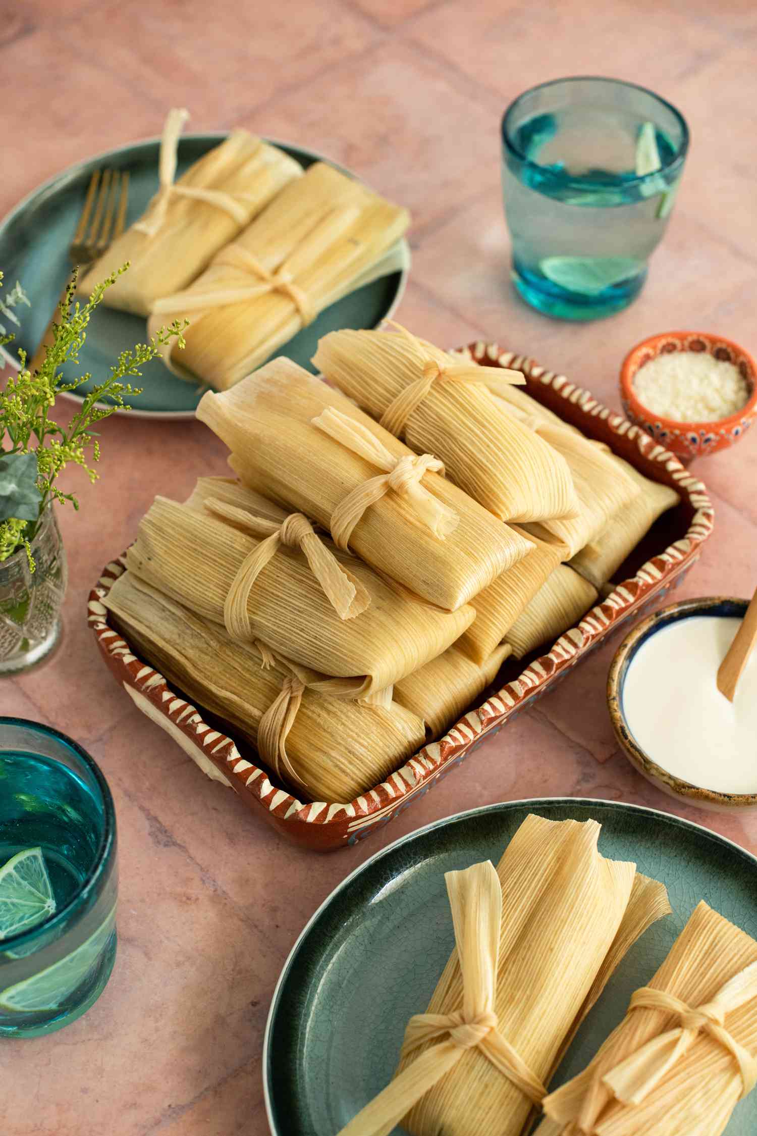 Platter of Chicken Tamales with Salsa Verde Surrounded by Two Plates with a Serving of Tamales and Small Bowls with Cotija Cheese, Crema, and Two Glasses with Lemon Water