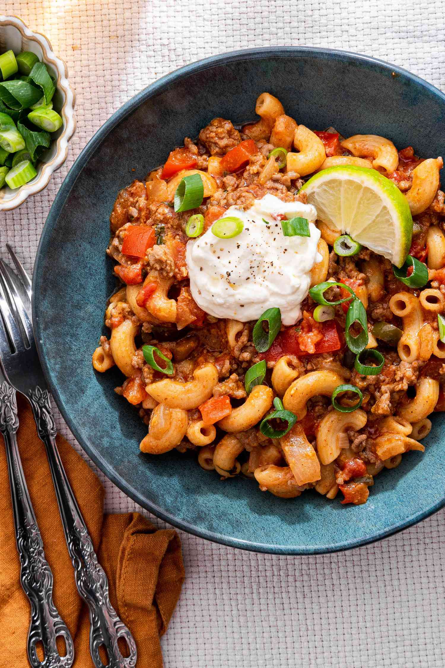 Bowl of One-Pot Taco Pasta Topped With Sour Cream, Sliced Scallions and a Lime Wedge, and in the Surroundings, a Bowl With Lime Wedges, a Bowl With Sliced Green Onions, a Bowl With Sour Cream, Glasses With a Drink, and a Mustard Colored Table Napkin With a Pair of Silverware