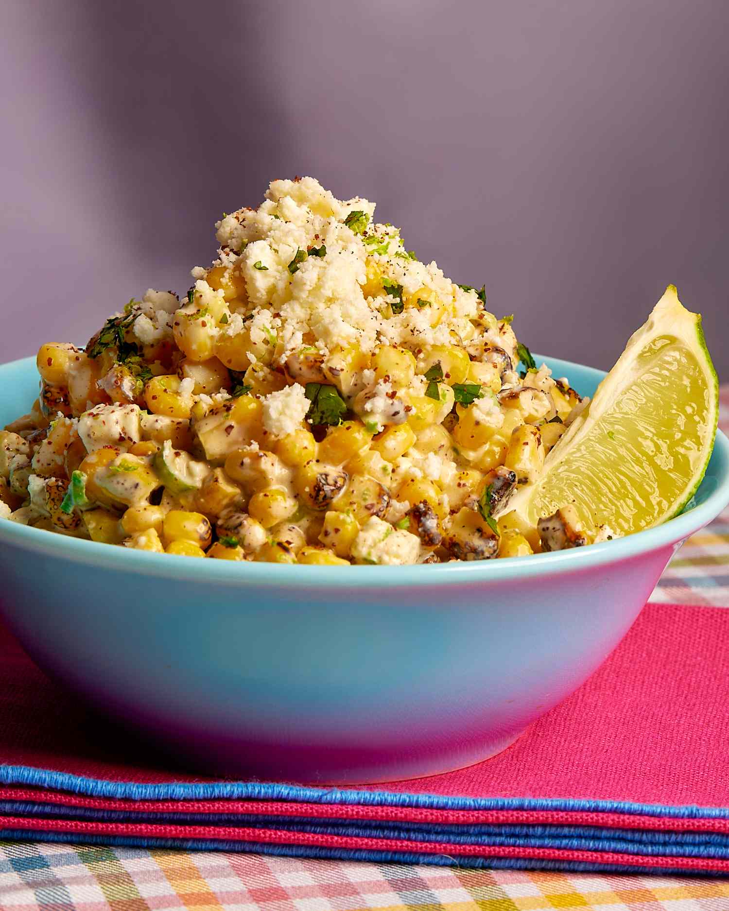 Creamy-looking corn kernels topped with crumbly cheese in a blue bowl with a lime wedge