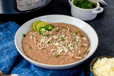 Pressure Cooker Refried Beans in a white bowl with lime wedges and sliced jalepenos nearby. The instant pot is in the background.