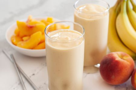 Two peach almond smoothies with whole bananas, peaches, and a plate of sliced peaches set around the smoothies.