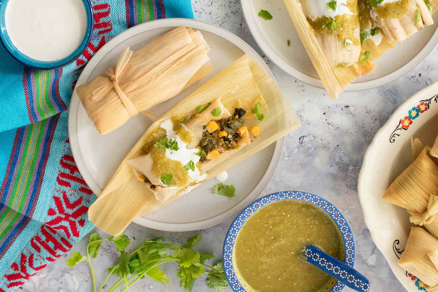 Poblano, Corn, and Cheese Tamales Topped with Salsa Verde, Crema, and Cilantro on Two Plates and Surrounded by a Bowl of Salsa Verde, a Bowl of Crema, and a Platter with More Tamales All on a Table Runner