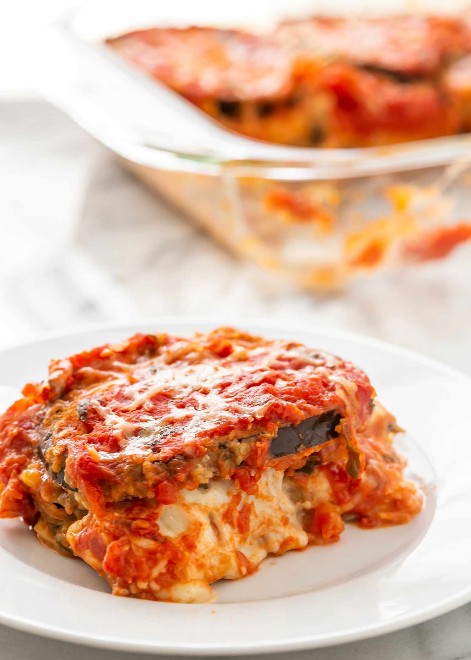 A slice of eggplant parmesan on a plate with the tray behind it.