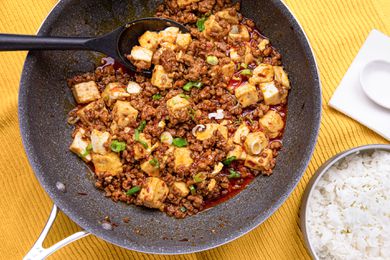 Mapo Tofu in a Pan With a Serving Spoon Next to a Bowl of Rice and a Soup Spoon on a Napkin, All on a Yellow Kitchen Towel
