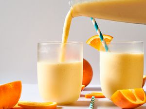 Two glasses of orange smoothie are served, with a blue-striped straw in one and orange slices on the rim and table
