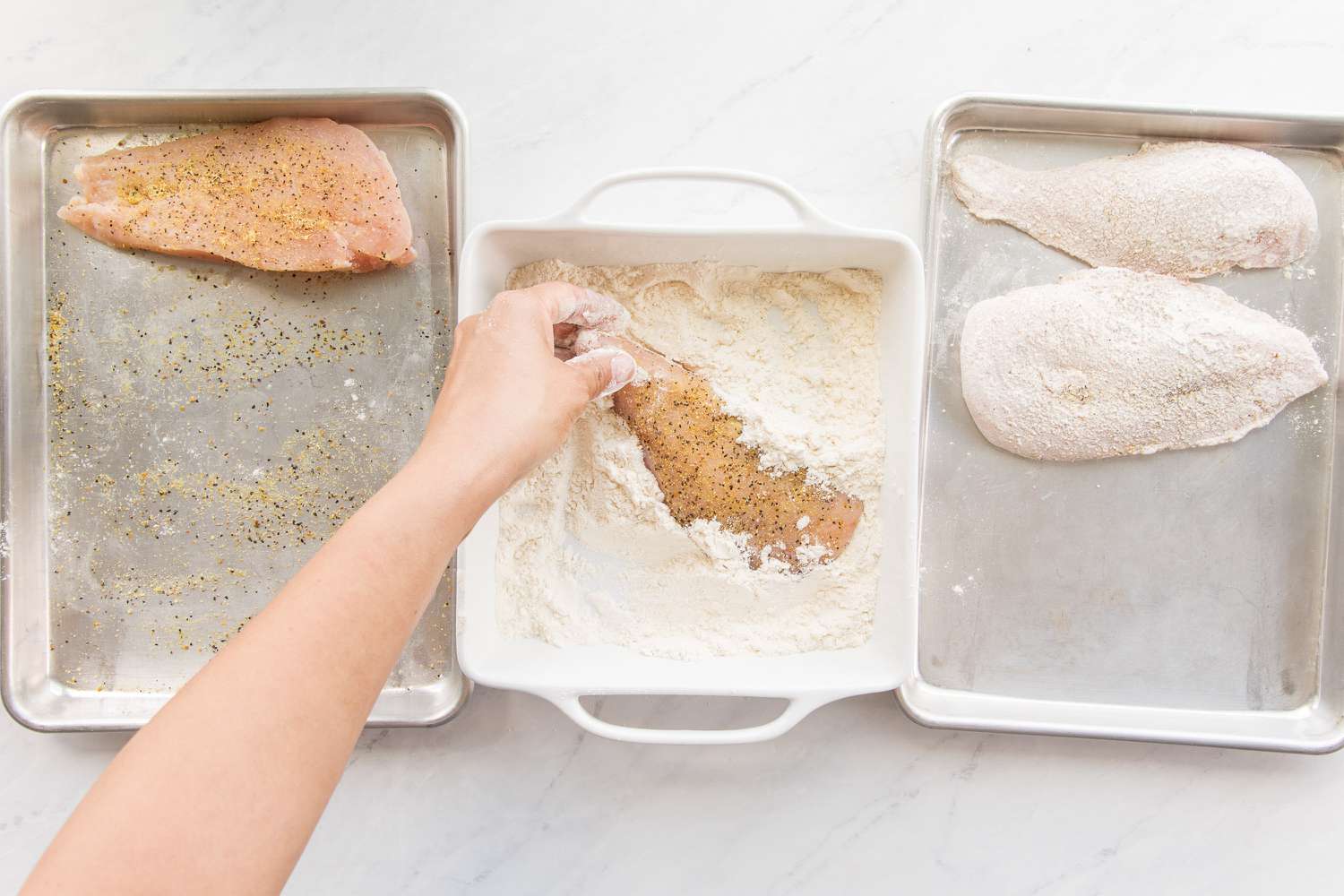 Dredging Station (L to R): Baking Pan with a Seasoned Chicken Cutlet, Casserole Dish with All-Purpose Flour and a Hand Covering a Cutlet in Flour, and a Baking Pan with Dredged Cutlets