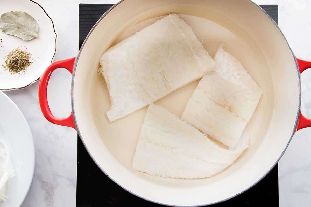 Three fillets of salt cod are at the bottom of a dutch oven. A scalloped plate has a bay leaf and ground spices to the left of the pot.