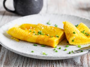 A Frenchstyle omelet garnished with chopped chives on a plate a fork to the side and a mug in the background