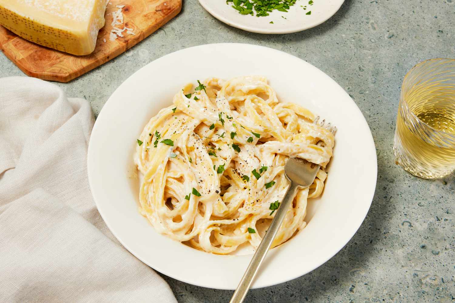Copycat Olive Garden alfredo in a bowl with a fork, and in the surroundings, a white table napkin, a small plate with minced herbs, a block of parmesan on a wooden board, and a glass with a drink