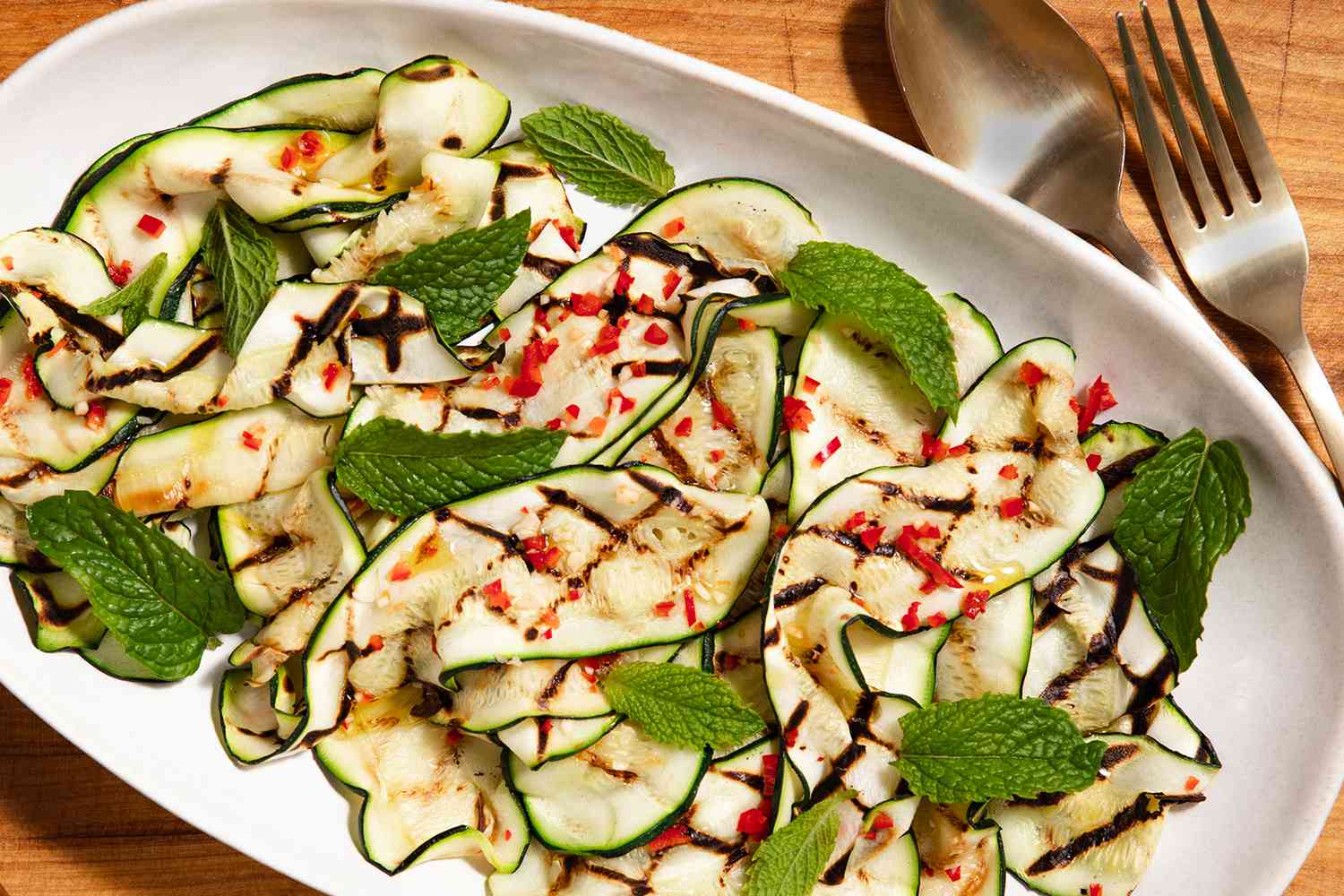 A plated grilled zucchini and squash salad with mint leaves and a fork and spoon alongside it